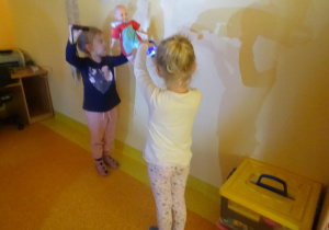 Dwie dziewczynki stoi przy ścianie, trzymają w jednej ręce zabawkę a w drugiej włączone latarki, obserwują powstające cienie na ścianie.